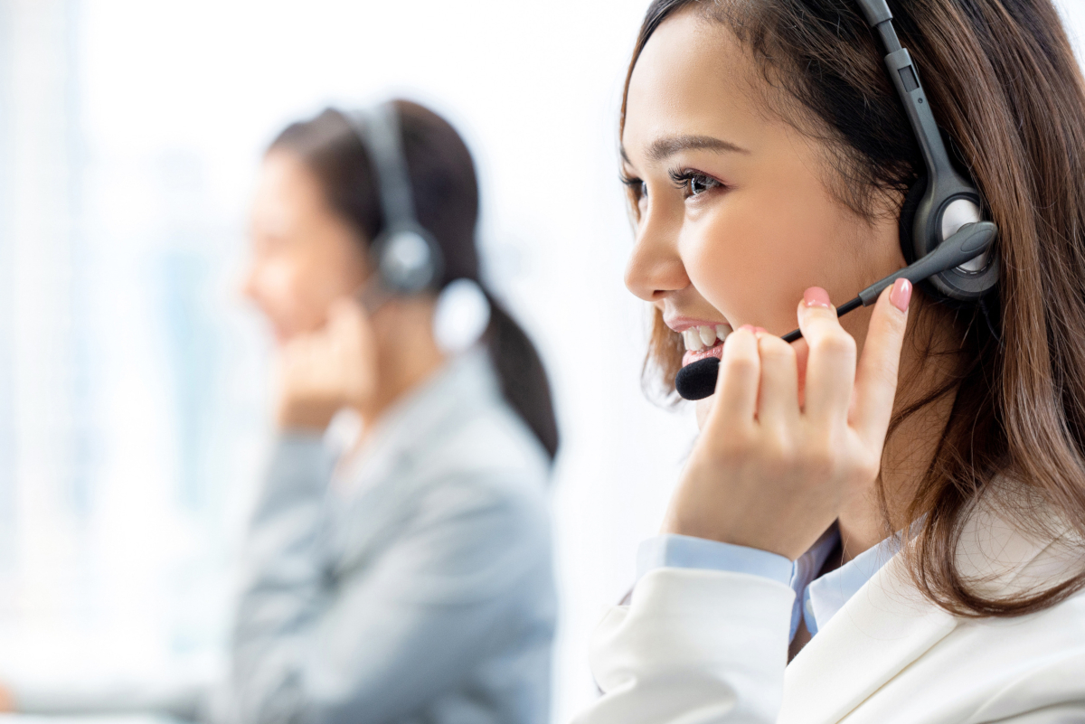 A close-up of a woman on a headset, with another woman on a headset in the background to represent contact centers.