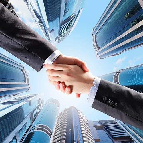 Low Angle View of Hands Shaking Across Backdrop of Blue Sky and Commercial Highrise Buildings, Partnership Concept