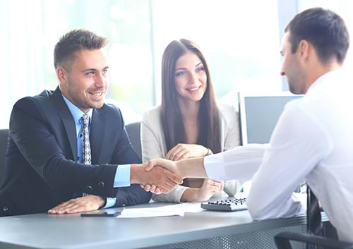 Man in White Shirt Shaking Hands with Smiling Man in Suit Across Table Seated Next to Smiling Businesswoman