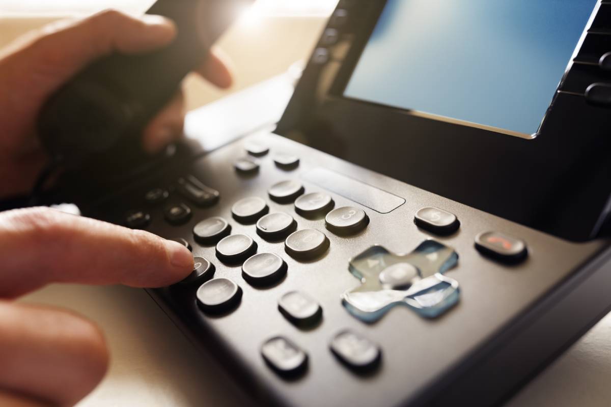 Close up of someone's hands typing in a number on a phone keypad, while holding it's receiver in the other hand in the background