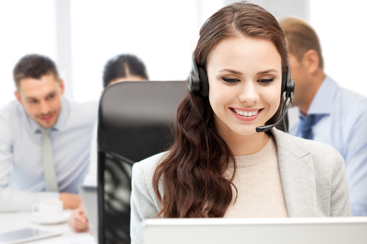 A smiling person working ontheir laptop while answering their desk phone through a headset