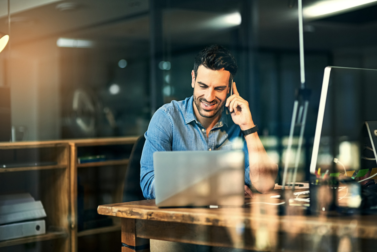 Man smiling while working on a computer and talking on a mobile phone