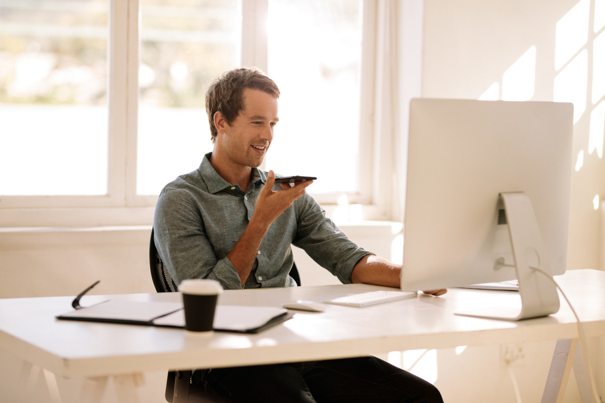 Man holding mobile phone in hand and talking on speaker looking at the computer.