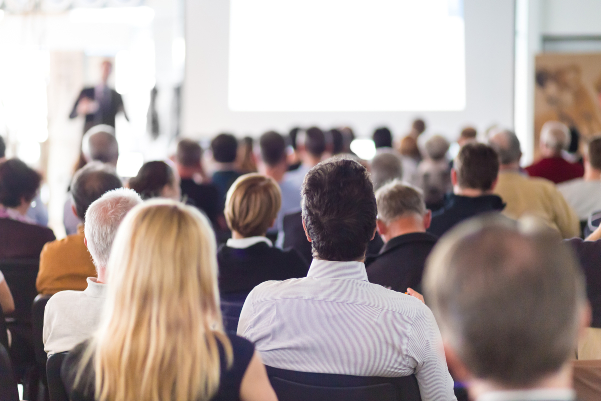 A crowd of seated people watching a speaker give a presentation at a convention similar to how Nova Communications will be speaking at TAG's Annual Convention.