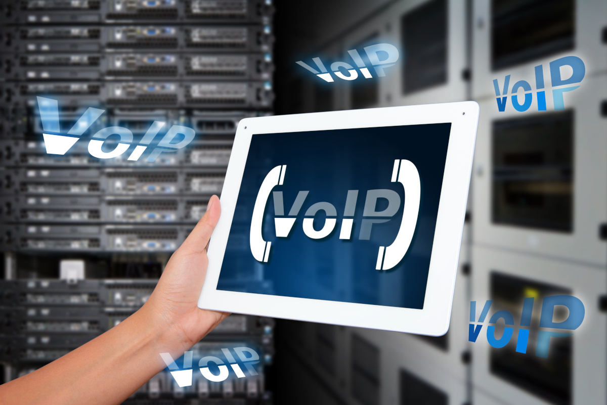 A tablet being held by one hand in a server room with the word VoIP brackted between two phone icons on it's screen, with the word 'VoIP' floating around the tablet.