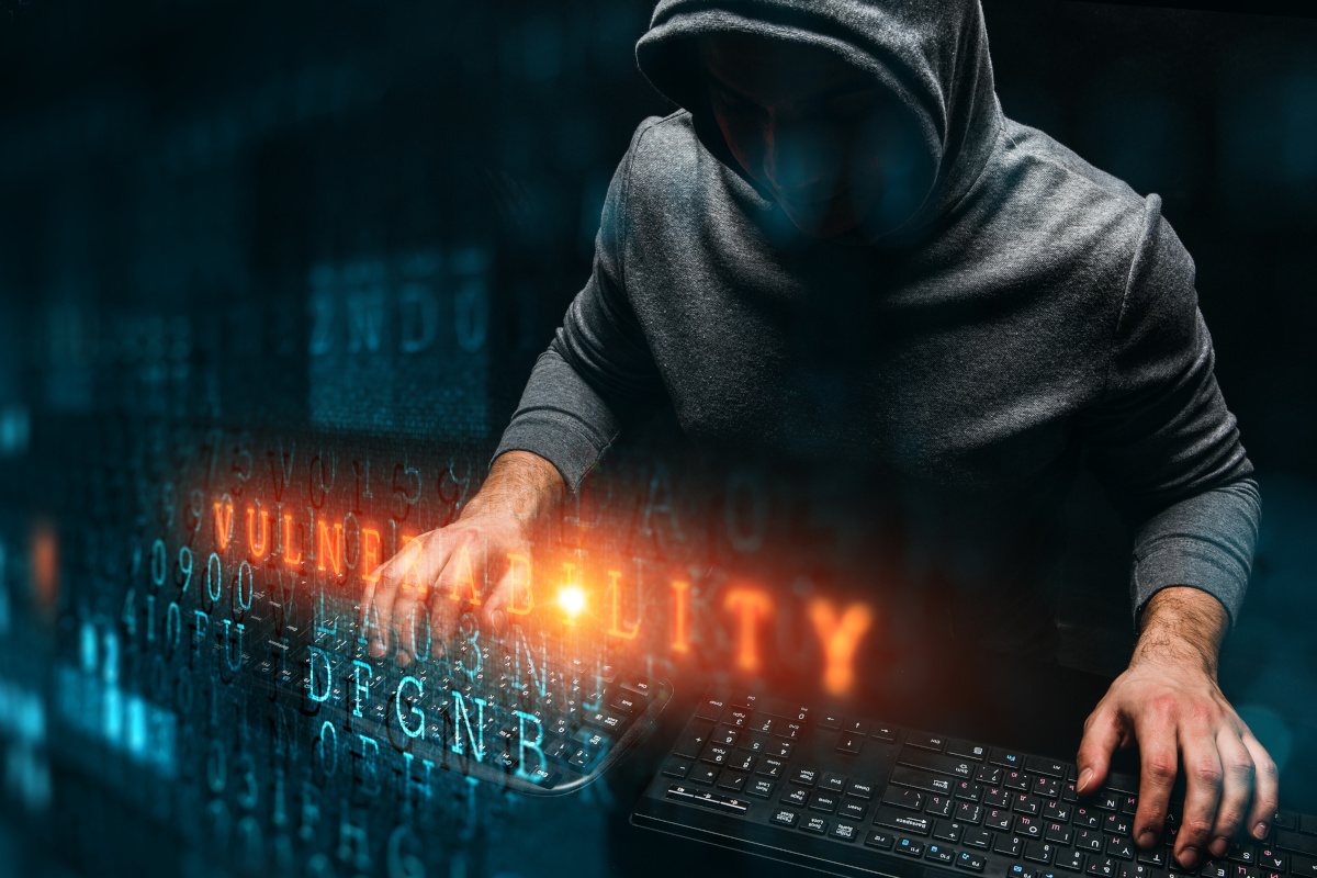 A hooded hacker typing on two keyboards with the word 'Vulnerability' in orange