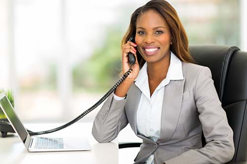 Smiling Woman in Suit Using Desk Phone to Call Customer Service/Support Seated in Front of Desk with Laptop Compter