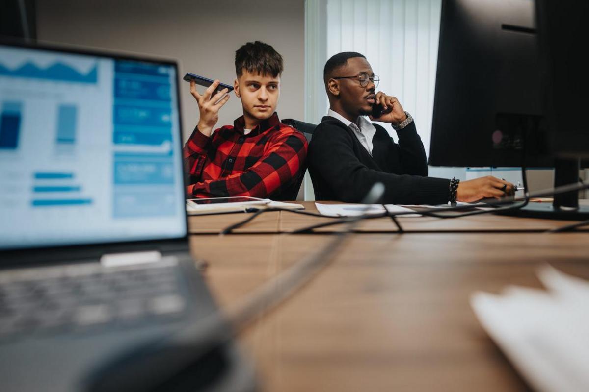 Two employees on business calls at a shared desk with laptops.