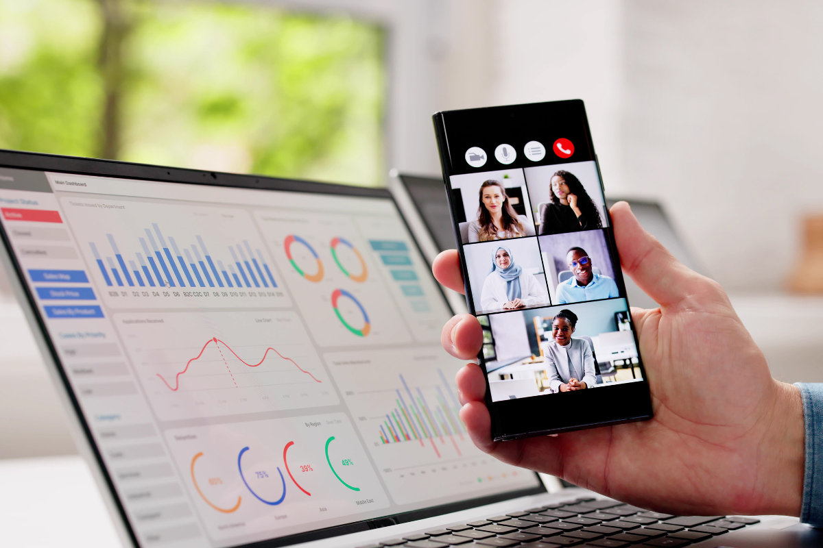 Video call on smart phone, with laptop behind showing charts.